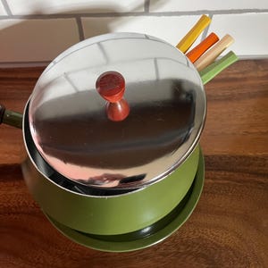 May include: A green fondue pot with a silver lid and a wooden knob. The pot is sitting on a black base and there are four colourful fondue forks sticking out of the lid.