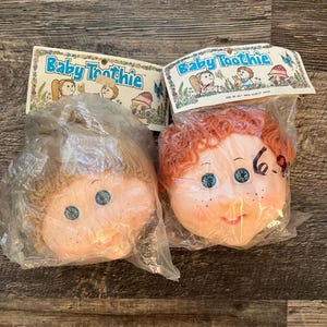 Op de afbeelding: Twee Baby Toothie poppenhoofden in plastic verpakking. Een poppenhoofd heeft bruin krullend haar en blauwe ogen. Het andere poppenhoofd heeft rood krullend haar en blauwe ogen. Beide poppenhoofden hebben sproeten en glimlachen.