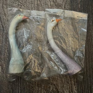 May include: Two white ceramic swan figurines with long necks and painted details. The swans are packaged in clear plastic bags.