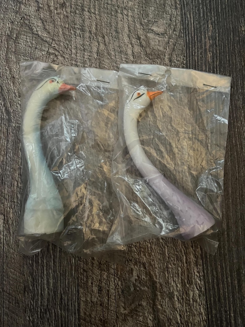 May include: Two white ceramic swan figurines with long necks and painted details. The swans are packaged in clear plastic bags.
