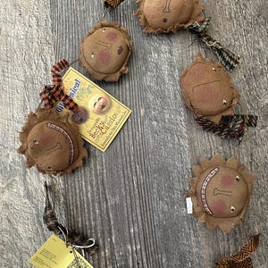 May include: A decorative garland featuring five stuffed brown fabric sun faces with stitched features and rosy cheeks. The garland includes patterned fabric ribbons and two attached paper tags with text including "Whimsical Folk Art" and "Designed By Becky Cassio".