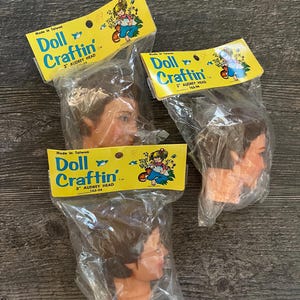 May include: Three doll heads in plastic packaging. Each package has a yellow label with the text "Doll Craftin'" and "3" Audrey Head". The doll heads are brown-haired with a side profile visible. The packages are on a dark wood surface.