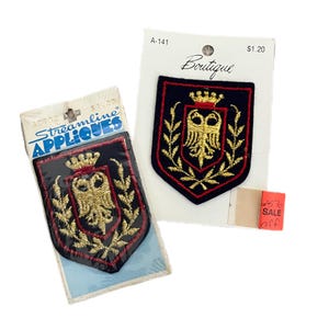 May include: Two embroidered shield-shaped patches featuring a gold double-headed eagle emblem topped with a crown and surrounded by gold laurel branches on a black background. The patches are attached to white retail display cards with text.