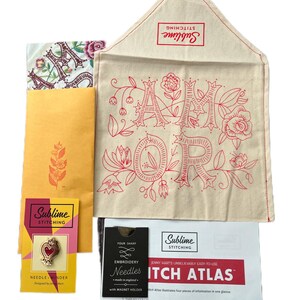 Op de afbeelding: Een set borduurbenodigdheden, waaronder een naaldhouder, borduurnaalden, een steekatlas en een stoffen tas met de woorden "Amor" geborduurd erop. De tas is beige met rode borduurwerk en bloemmotieven.