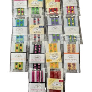 May include: A collection of packaged ribbons in various colors and patterns. Designs include polka dots, stripes, and gingham. The ribbons are labeled with the terms "polka dot organdy", "striped grosgrain", "accent organdy", and "gingham".