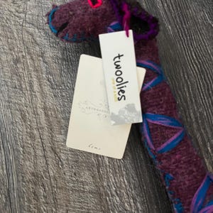 Pode incluir: Um brinquedo de pel&uacute;cia de girafa roxa feito &agrave; m&atilde;o com detalhes em azul e rosa. A girafa tem um pesco&ccedil;o longo e uma etiqueta que diz "twooolies one of a kind". O brinquedo de pel&uacute;cia &eacute; feito de um material macio e texturizado e &eacute; adequado para crian&ccedil;as.