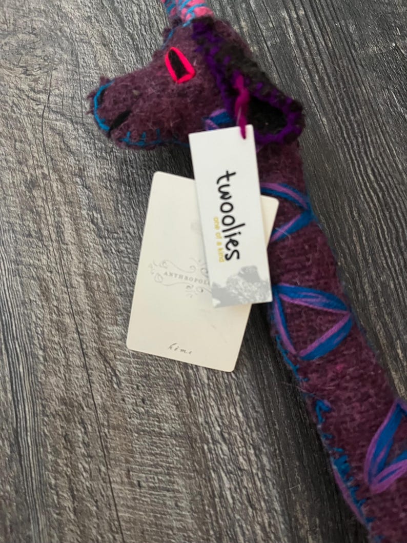 Pode incluir: Um brinquedo de pel&uacute;cia de girafa roxa feito &agrave; m&atilde;o com detalhes em azul e rosa. A girafa tem um pesco&ccedil;o longo e uma etiqueta que diz "twooolies one of a kind". O brinquedo de pel&uacute;cia &eacute; feito de um material macio e texturizado e &eacute; adequado para crian&ccedil;as.