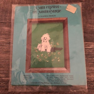 May include: A vintage "Mini Crewel" embroidery kit by NeedleMagic, featuring a hand-painted design of a white dog with butterflies on a green background. The kit includes cotton fabric, wool yarn, a needle, and instructions. The artwork is framed and measures 12.7cm x 17.8cm.