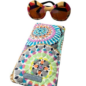 Könnte beinhalten: Eine übergroße, runde Sonnenbrille mit einem farbenfrohen geometrischen Muster und braunen Gläsern, zusammen mit einem gesteppten Brillenetui von Vera Bradley mit einem bunten Blumenmuster.