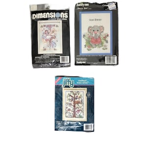May include: Three cross-stitch kits are displayed. The top left kit is from Dimensions, featuring a Victorian carousel scene. The top right kit from Janlynn has a koala bear with the text "God Bless!". The bottom kit from Jiffy shows a tree scene.