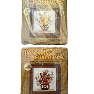 May include: Two vintage "Needle Pointers" cross-stitch kits. Each kit features a framed floral design. One depicts yellow flowers, the other a rust and brown bouquet. The kits are packaged in clear plastic with a burlap-textured background.