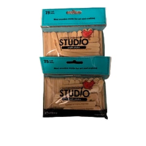 Pode incluir: Duas embalagens de mini palitos de madeira para artesanato. Cada embalagem tem um rótulo turquesa com o texto "75" e "Mini palitos de madeira para arte e artesanato". A marca "STUDIO" está impressa no rótulo com um logotipo de pássaro vermelho.