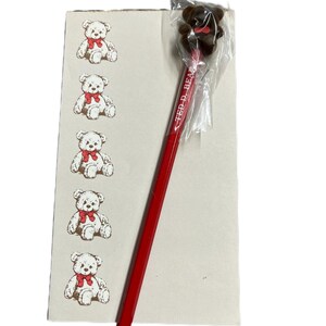 May include: A red pencil with the text "TED D. BEAR" and a brown bear topper. A small notepad with six illustrations of white teddy bears wearing red bows is also present. The pencil is from Taiwan.