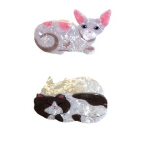 May include: Two cat-shaped hair clips. The top clip is a white cat with pink ears and spots. The bottom clip is a sleeping cat with brown and white markings. Both clips have a pearlescent finish.