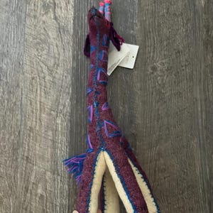 Pode incluir: Um brinquedo de pel&uacute;cia de girafa feito &agrave; m&atilde;o em tons de roxo, azul e creme. A girafa tem um pesco&ccedil;o longo com desenhos geom&eacute;tricos azuis e roxos. O brinquedo tem uma barriga e pernas cor de creme. As orelhas e a crina da girafa s&atilde;o feitas de fio azul e roxo.