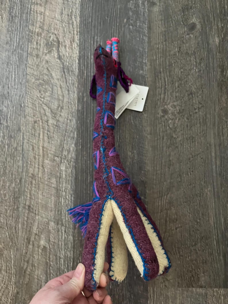 Pode incluir: Um brinquedo de pel&uacute;cia de girafa feito &agrave; m&atilde;o em tons de roxo, azul e creme. A girafa tem um pesco&ccedil;o longo com desenhos geom&eacute;tricos azuis e roxos. O brinquedo tem uma barriga e pernas cor de creme. As orelhas e a crina da girafa s&atilde;o feitas de fio azul e roxo.