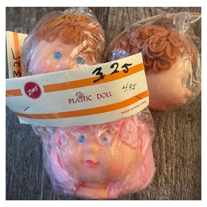 Op de afbeelding: Drie plastic poppenhoofden met garenhaar. Twee hoofden hebben bruin garenhaar en één hoofd heeft roze garenhaar. De poppenhoofden zijn verpakt in plastic zakken met een prijskaartje waarop "3.25" en "4.95" staat. Het prijskaartje vermeldt ook "Plastic Doll" en "Zims".