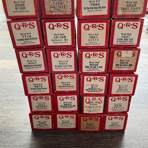 May include: A collection of vintage Q.R.S. piano rolls in red boxes. Each box features song titles and artist information. The boxes are stacked, showcasing a variety of musical selections from different eras. The text on the boxes is in black and white.