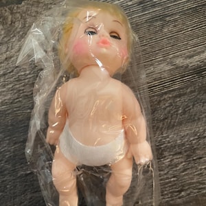 Puede incluir: Una muñeca bebé vintage con cabello rubio, mejillas rosadas y ojos cerrados, empaquetada en plástico transparente. La muñeca lleva un pañal blanco. Los brazos y las piernas de la muñeca están ligeramente doblados.