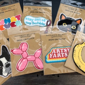 Könnte beinhalten: Ein Set mit zehn Bügelbildern mit verschiedenen Designs, darunter eine schwarze Katze, eine Banane, ein Ballontier und ein Hund. Die Patches sind alle auf braunem Hintergrund mit dem Text "SMARTY PANTS PAPER CO. IRON ON PATCH" auf jedem gedruckt.