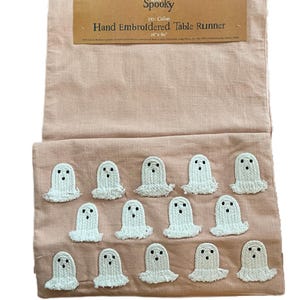 May include: A peach-colored, hand-embroidered table runner with white ghost appliqués. The runner is 16" x 90" and made of 100% cotton. The tag reads "Spooky" and "Hand Embroidered Table Runner".