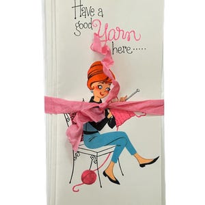 May include: A stack of blank greeting cards with a pink ribbon tied around them. The front of the card features a cartoon illustration of a woman knitting with the text "Have a good yarn here..."