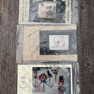 May include: Three different craft patterns featuring cats. The first pattern is for a pillow with the text "Have a Purrfect Day". The second pattern is for a cross stitch with the text "My Cat is an Angel". The third pattern is for five noteholders with the text "You are Special Patterns" and "#255 Prim Noteholders".