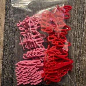 May include: A clear plastic bag filled with pink and red plastic cake toppers. The toppers are in the shape of the words "Happy Birthday" and "Thank You" in a cursive font. The bag is on a wooden surface.