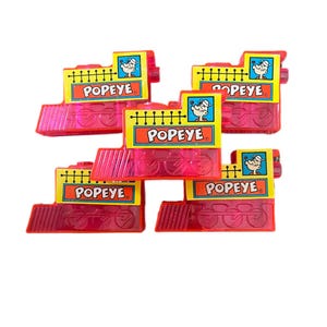 May include: Several pink toy train cars with the word "POPEYE" on a yellow sign. Each train car has a picture of Popeye the Sailor Man. The train cars are made of a translucent pink plastic.