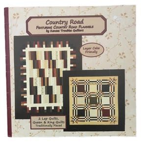 Könnte beinhalten: Ein Quiltmusterbuch mit dem Titel "Country Road" mit zwei Quilt-Designs. Das Cover zeigt zwei Quiltblock-Beispiele in neutralen Tönen. Der Text lautet "Featuring Country Road Flannels by Kansas Troubles Quilters".
