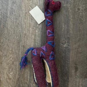 Pode incluir: Um brinquedo de girafa de pel&uacute;cia feito &agrave; m&atilde;o em tons de roxo, azul e rosa. A girafa tem um pesco&ccedil;o longo com detalhes triangulares azuis e roxos, um chifre rosa e uma cauda tran&ccedil;ada. Uma etiqueta branca est&aacute; anexada.