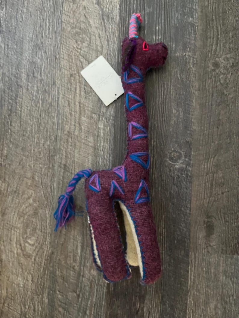 Pode incluir: Um brinquedo de girafa de pel&uacute;cia feito &agrave; m&atilde;o em tons de roxo, azul e rosa. A girafa tem um pesco&ccedil;o longo com detalhes triangulares azuis e roxos, um chifre rosa e uma cauda tran&ccedil;ada. Uma etiqueta branca est&aacute; anexada.