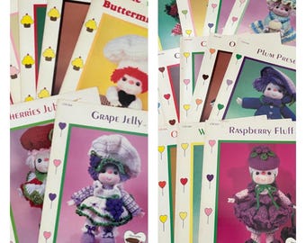 May include: A collection of vintage crochet pattern booklets featuring whimsical doll designs. The covers showcase colorful illustrations of the dolls, each with a unique outfit and name, such as "Grape Jelly" and "Raspberry Fluff". The booklets are stacked, displaying their titles.