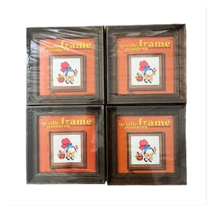 Peut inclure: Quatre cadres carrés en bois foncé pour point de croix emballés dans du plastique transparent. Chaque cadre présente une carte orange avec le texte needle frame pointers et une illustration d'un enfant en tenue bleue tenant un panier de fleurs.