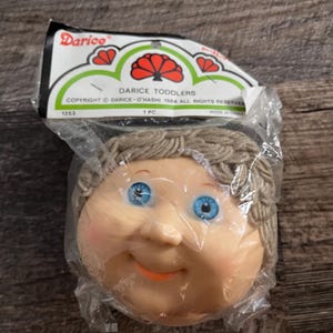 Op de afbeelding: Een Darice Toddlers poppenhoofd in een plastic verpakking. Het poppenhoofd heeft blauwe ogen, een glimlachende uitdrukking en lichtbruin garen haar. De verpakking bevat het Darice-logo en tekst.
