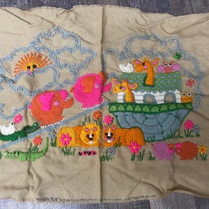 May include: A beige fabric wall hanging with embroidered animals, including a pink elephant, a giraffe, a lion, and a crocodile. The animals are on a blue and green boat with a white roof. The background is a light blue sky with white clouds.