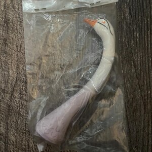 May include: A white swan-shaped plastic whistle with an orange beak and black eyes. The whistle is packaged in a clear plastic bag.