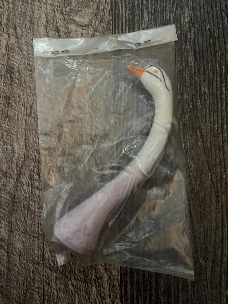May include: A white swan-shaped plastic whistle with an orange beak and black eyes. The whistle is packaged in a clear plastic bag.