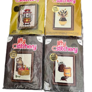 May include: Four vintage Jiffy Stitchery cross-stitch kits with a brown background and a white frame. The kits feature images of a cable car, cattails and wheat, cider, and coffee. Each kit includes a frame offer inside.