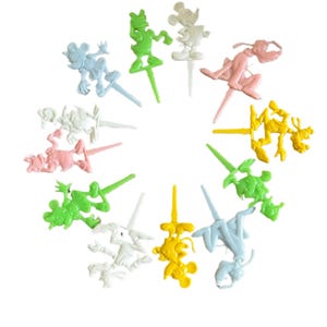 Puede incluir: Una colección de palillos de comida de personajes de plástico de colores. Los palillos presentan varios personajes de dibujos animados en tonos de blanco, rosa, amarillo, verde y azul. Cada palillo tiene un extremo puntiagudo para insertar en los alimentos.
