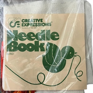 May include: A sealed craft kit labelled "Needle Book" with a bird design. The kit includes a tan paper cover with green text and a bird graphic, along with a bundle of colourful yarn and a piece of off-white fabric.
