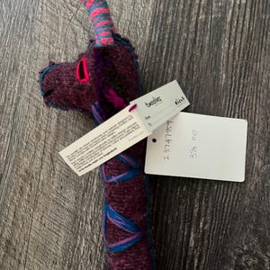 Pode incluir: Uma girafa de pel&uacute;cia feita &agrave; m&atilde;o com corpo roxo, detalhes azuis e rosa e uma etiqueta. A cabe&ccedil;a da girafa apresenta uma boca rosa e costuras azuis. A etiqueta diz "twoolies".