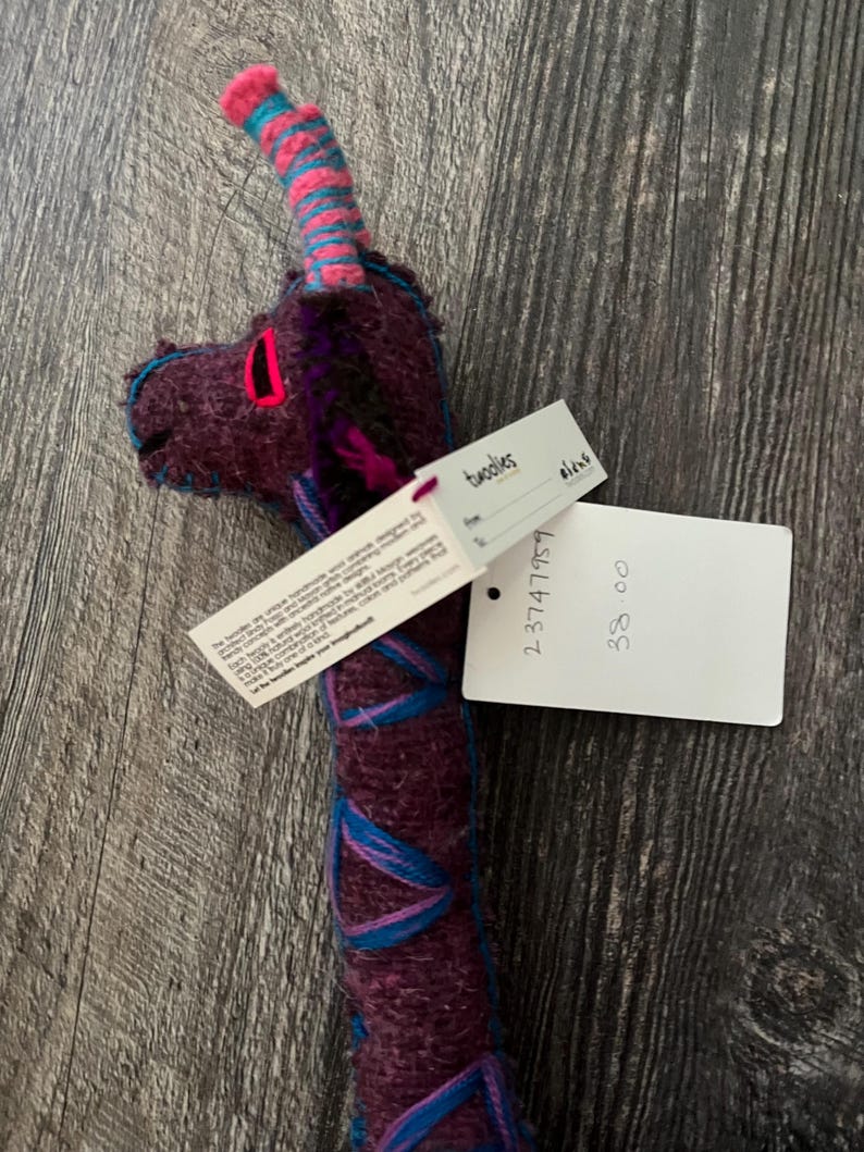 Pode incluir: Uma girafa de pel&uacute;cia feita &agrave; m&atilde;o com corpo roxo, detalhes azuis e rosa e uma etiqueta. A cabe&ccedil;a da girafa apresenta uma boca rosa e costuras azuis. A etiqueta diz "twoolies".