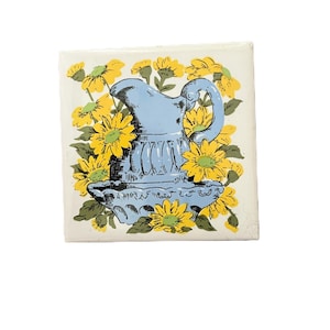 Puede incluir: Azulejo de cerámica cuadrado con una jarra azul claro rodeada de girasoles amarillos con hojas verdes. La jarra tiene un asa decorativa y un diseño detallado. El fondo es blanquecino.