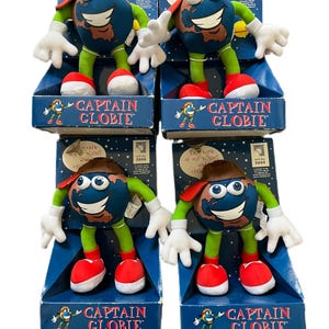 Peut inclure: Quatre poupées Captain Globie en peluche dans leurs boîtes d'origine. Chaque poupée est un globe souriant avec des gants blancs, des bras et des jambes verts, des chaussures rouges et un chapeau marron. Les boîtes sont bleues avec le texte "CAPTAIN GLOBIE".