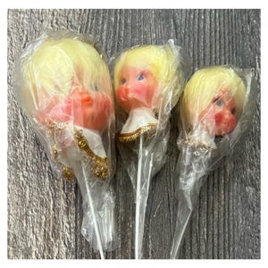 以下が含まれることがあります： 透明なプラスチックに包まれた、ブロンドの髪とバラ色の頬を持つ、3つのヴィンテージ人形の頭部。人形は青い目を持ち、白と金の装飾的な襟を着用しています。頭のサイズは様々です。