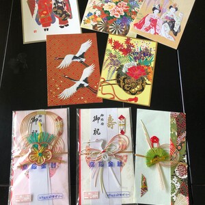 Op de afbeelding: Een verzameling Japanse wenskaarten en enveloppen. De kaarten tonen illustraties van geisha's, bloemen en kranen. De enveloppen hebben gouden en zilveren draadaccenten en Japanse tekst.