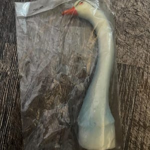 May include: A white plastic goose decoy with a red beak and black eyes. The decoy is in a clear plastic bag.