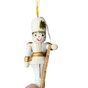 May include: A small, white wooden toy soldier ornament with gold accents. The ornament has a smiling face, a tall hat, and a wooden staff. A gold string is attached for hanging. The ornament is likely for Christmas or holiday decor.
