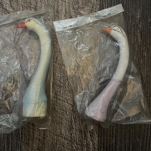 May include: Two ceramic swan figurines in individual plastic packaging. The swan on the left is light blue and white, while the swan on the right is purple and white.